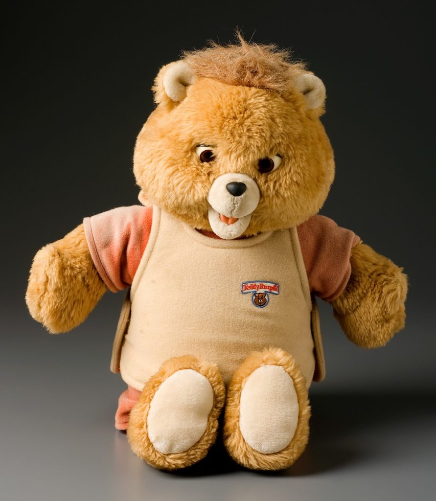 Animatronic toy teddy bear with plush fur body, large head, small ears and moveable mouth. The bear is light brown in colour and wears a short sleeved orange top beneath a tan coloured tunic. The tunic has an embroidered logo with the bear's name 'Teddy Ruxpin'. The body of the bear contains a cassette tape player which can be accessed by lifting the tunic. The inserted audio cassettes synchronise the talking apparatus which control the bear's mouth with the story heard on the tape.
